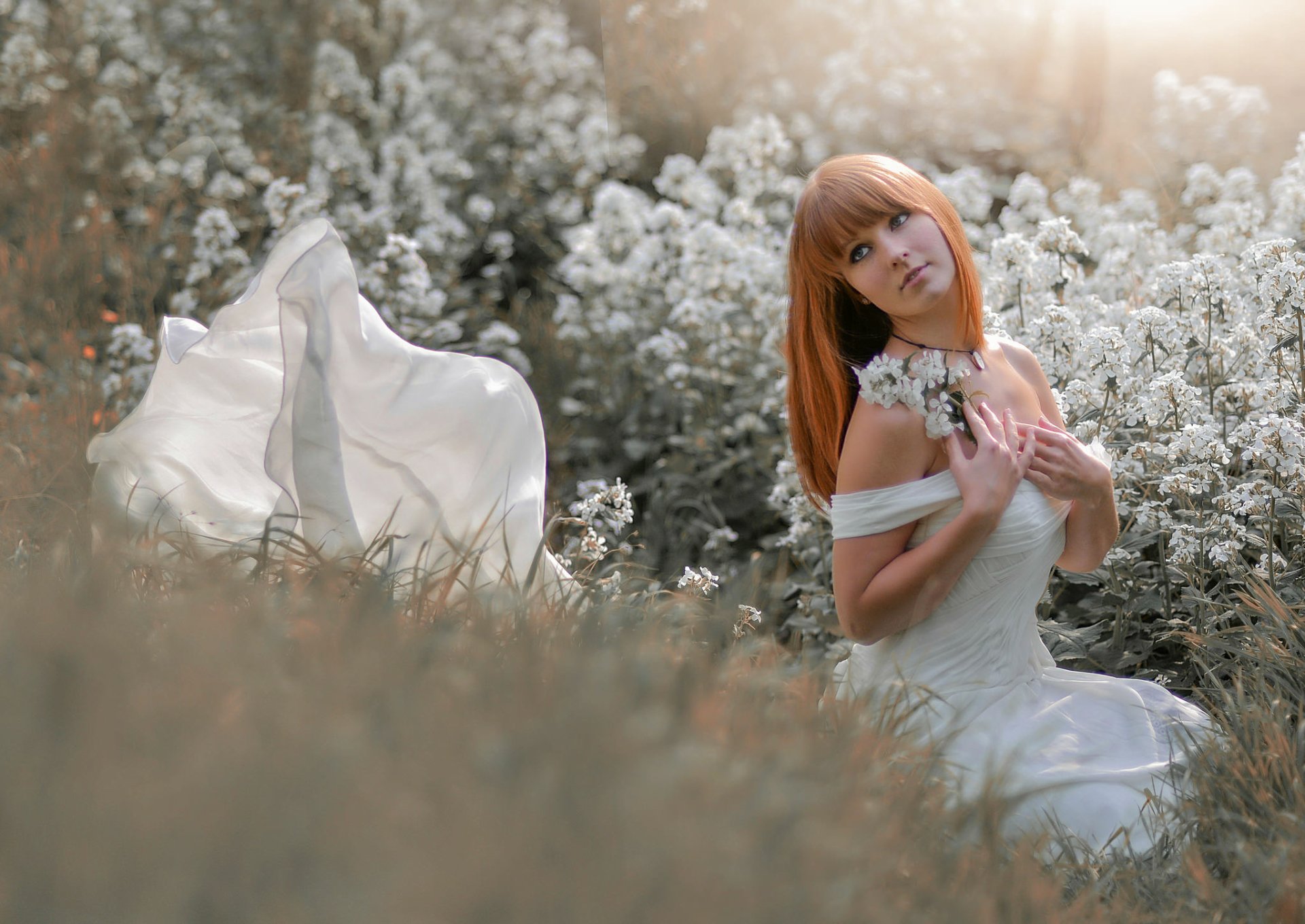 A serene outdoor scene featuring a redhead bride in a stunning wedding dress, surrounded by blooming white flowers, creating a romantic and dreamy atmosphere. HD wallpaper.