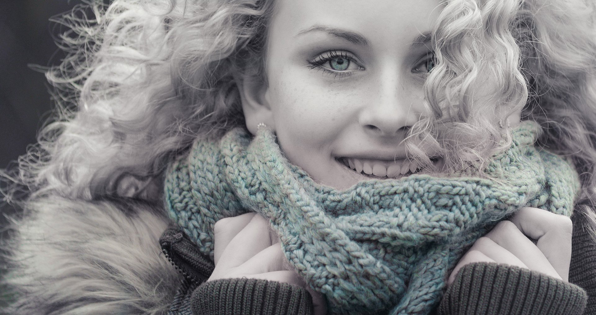 A smiling woman with blonde curly hair and blue eyes, wrapped in a cozy scarf, radiates warmth and charm in this HD desktop wallpaper.