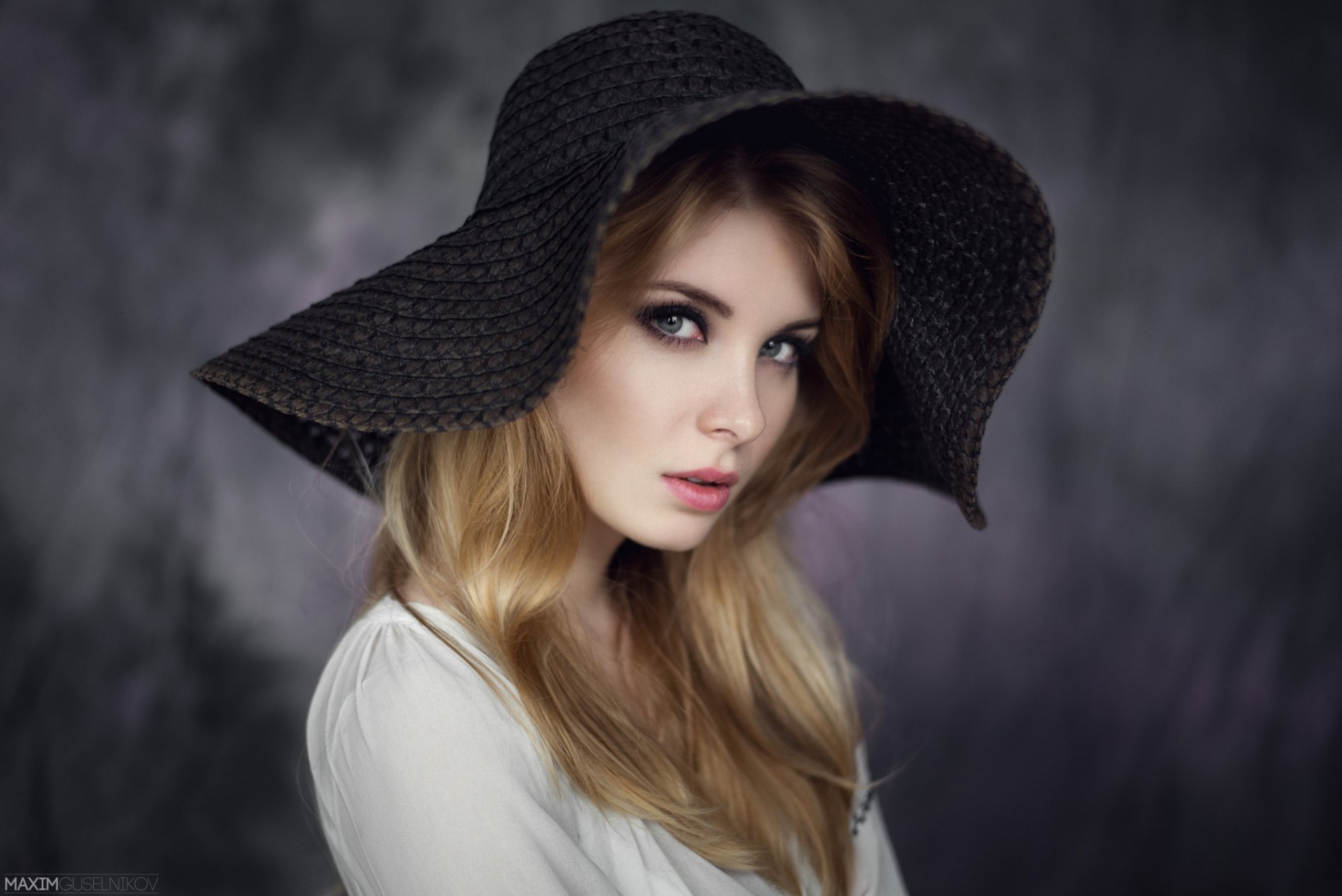 A woman with blonde hair and blue eyes wears a large black hat, gazing softly at the camera. The background features a bokeh effect, enhancing the portrait's elegance.