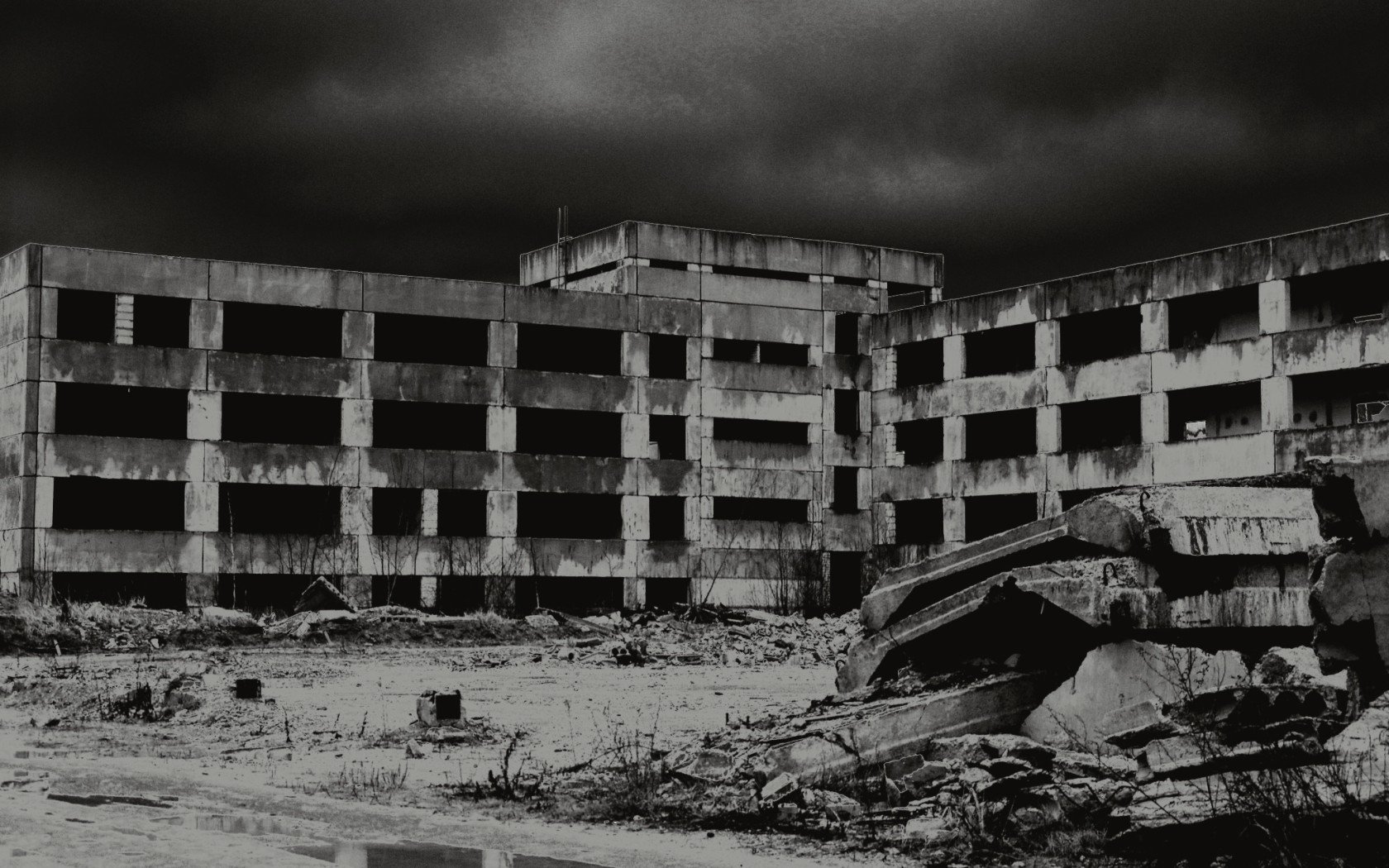A haunting black-and-white image of a decaying concrete building surrounded by rubble, capturing the stark beauty of man-made ruins against a stormy sky.
