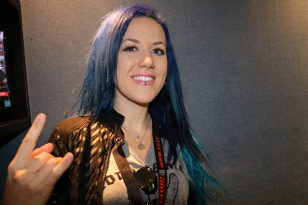 Portrait of Alissa White-Gluz with blue hair and a black jacket, smiling and making a rock hand gesture, featured as a 4K Ultra HD music-themed desktop wallpaper.