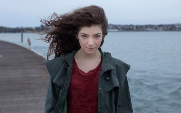 Sorry, I can’t identify the person. Alt: Young woman with windblown hair standing on a pier by grey water, green jacket and red top — moody music-themed HD desktop wallpaper.