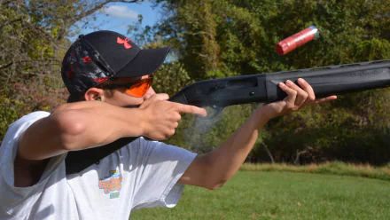 Man-made shotgun being fired by a man outdoors, captured in 4K Ultra HD quality, with a red shotgun shell ejecting mid-air against a green, natural background.