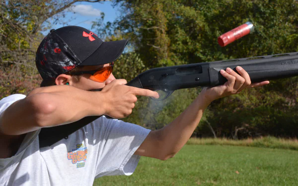 Man-made shotgun being fired by a man outdoors, captured in 4K Ultra HD quality, with a red shotgun shell ejecting mid-air against a green, natural background.