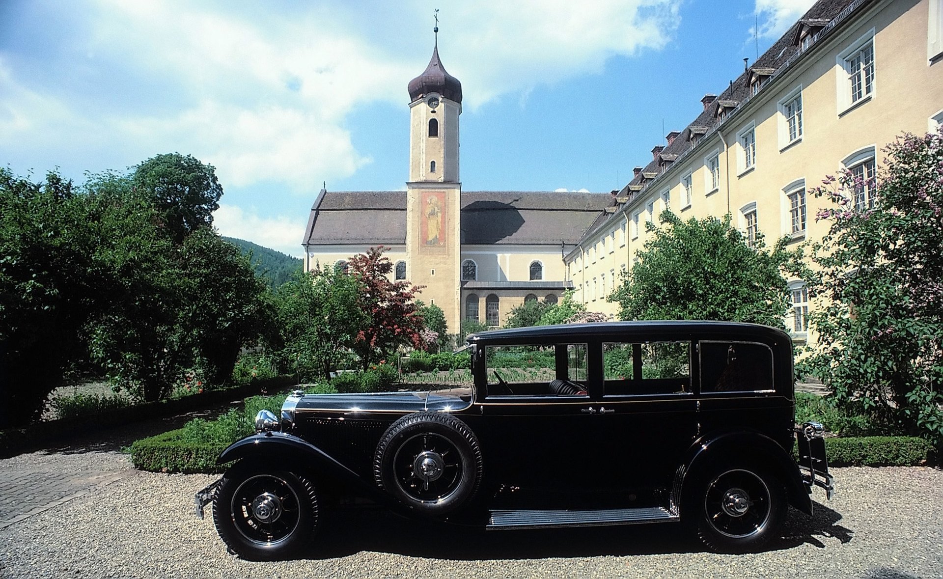 HD PC desktop wallpaper: black Mercedes‑Benz W08 parked in a sunlit European courtyard with a church tower and row of yellow buildings beneath a blue sky.