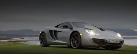 Silver McLaren MP4-12C parked on grass under an overcast sky, headlights on; 2K Quad HD PC desktop wallpaper and background showcasing the sports car's profile.
