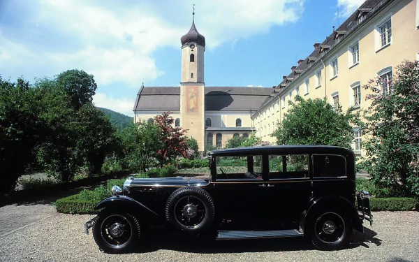 HD PC desktop wallpaper: black Mercedes‑Benz W08 parked in a sunlit European courtyard with a church tower and row of yellow buildings beneath a blue sky.