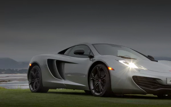 Silver McLaren MP4-12C parked on grass under an overcast sky, headlights on; 2K Quad HD PC desktop wallpaper and background showcasing the sports car's profile.