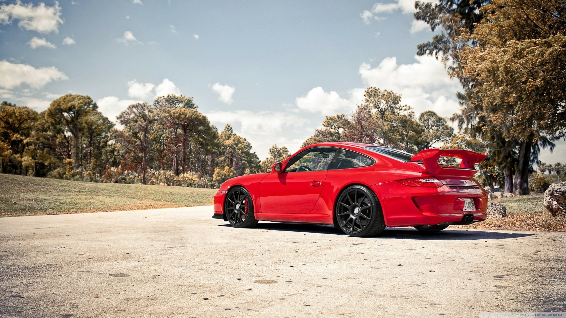 A red Porsche 911 GT3 parked on a paved road surrounded by trees under a partly cloudy sky, featured as an HD PC desktop wallpaper and background.