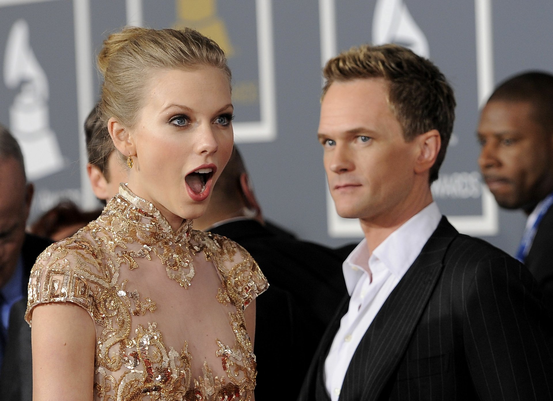 Taylor Swift, a renowned American singer with blonde hair, is captured in a candid moment with Neil Patrick Harris at a glamorous event, showcasing their expressive reactions.