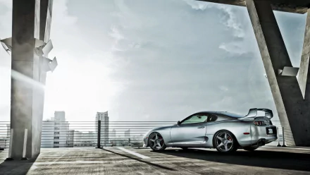 HD desktop wallpaper featuring a silver Toyota Supra parked on an urban rooftop with a cityscape and cloudy sky in the background.
