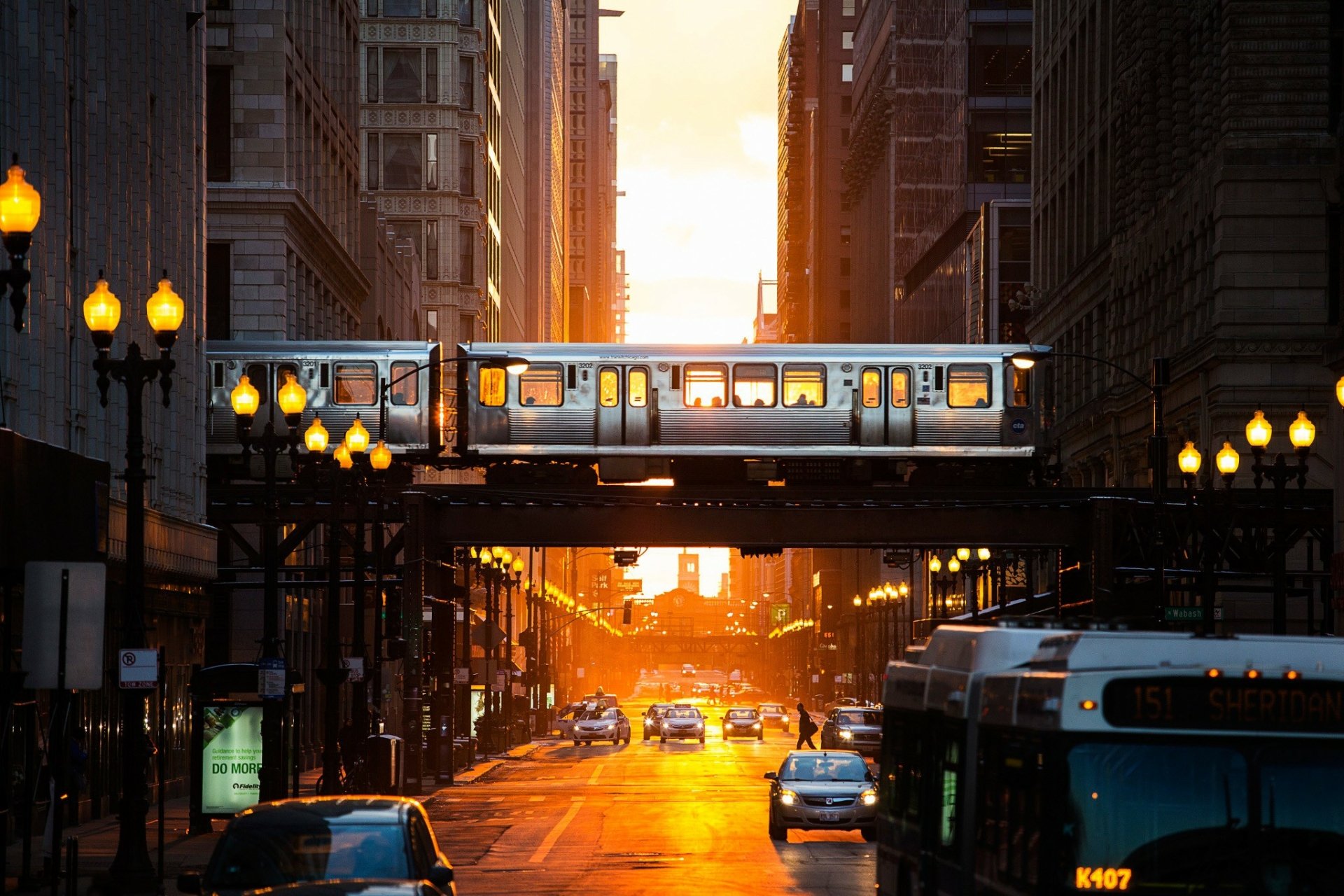 A vibrant Chicago scene at sunset, featuring a train elevated above a bustling street, illuminated by golden light and city lamps. A captivating urban wallpaper for a PC desktop.