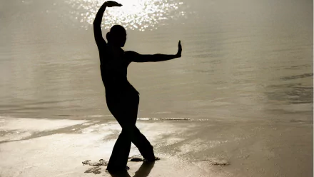 A silhouette of a woman practicing yoga by the water, with sunlight reflecting on the surface, creating a tranquil beach scene. This HD desktop wallpaper enhances any background.