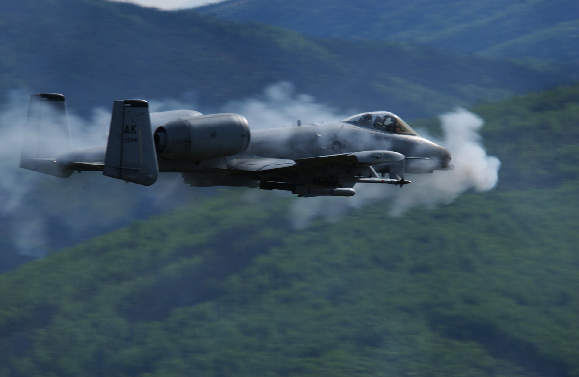 HD desktop wallpaper of a Fairchild Republic A-10 Thunderbolt II military aircraft flying low over green mountainous terrain with smoke trailing behind.