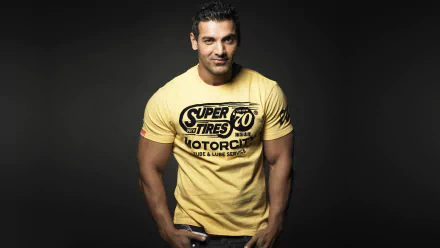 HD desktop wallpaper featuring celebrity John Abraham in a yellow graphic tee against a dark background.