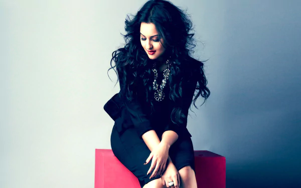 HD wallpaper featuring Sonakshi Sinha, showcasing her stylish look with dark, wavy hair and an elegant outfit, exuding confidence and charm against a muted background.