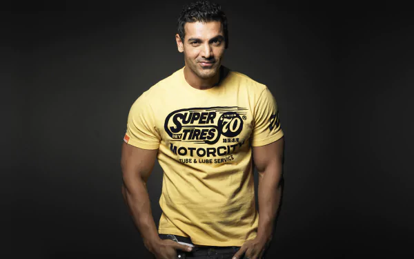 HD desktop wallpaper featuring celebrity John Abraham in a yellow graphic tee against a dark background.