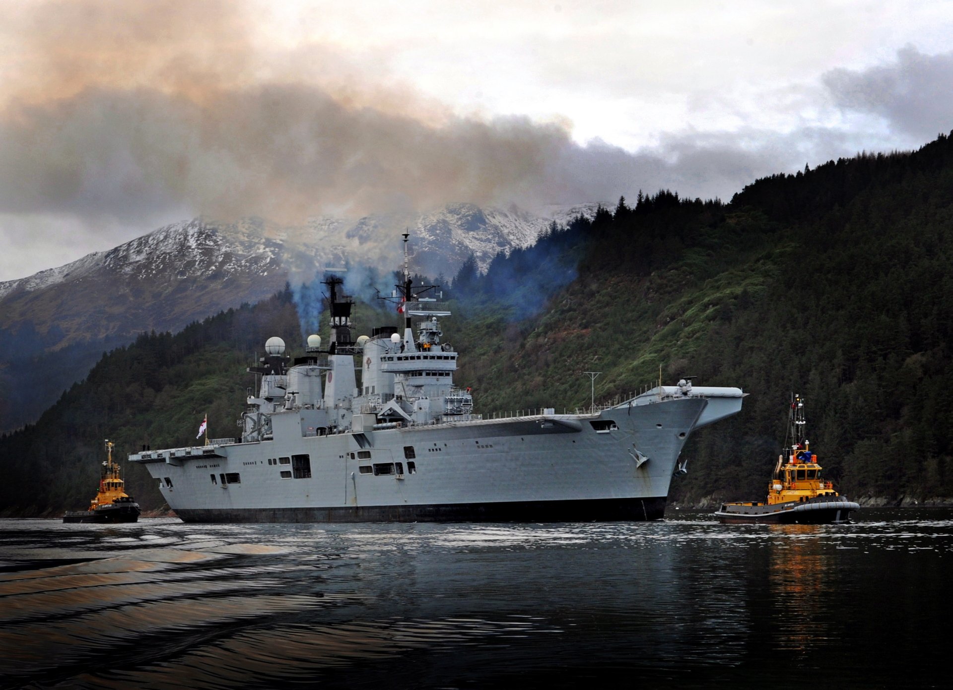 HD wallpaper of the HMS Illustrious (R06) aircraft carrier navigating calm waters, flanked by tugboats with mountainous terrain and overcast skies in the background.
