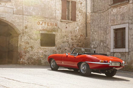 A red Jaguar E-Type convertible parked on a sunlit cobblestone street against an old stone building, captured in stunning 4K Ultra HD for a PC desktop wallpaper.