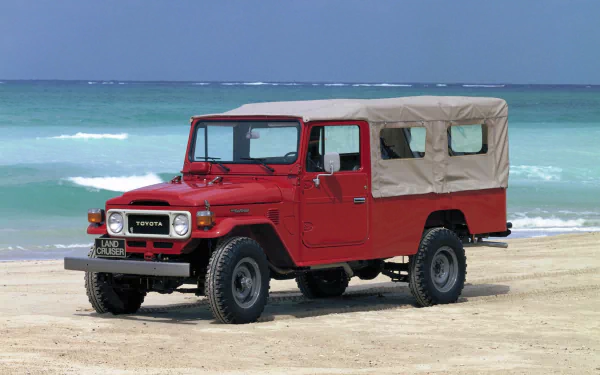 HD PC desktop wallpaper featuring a red Toyota Land Cruiser parked on a sandy beach with the ocean and blue sky in the background.