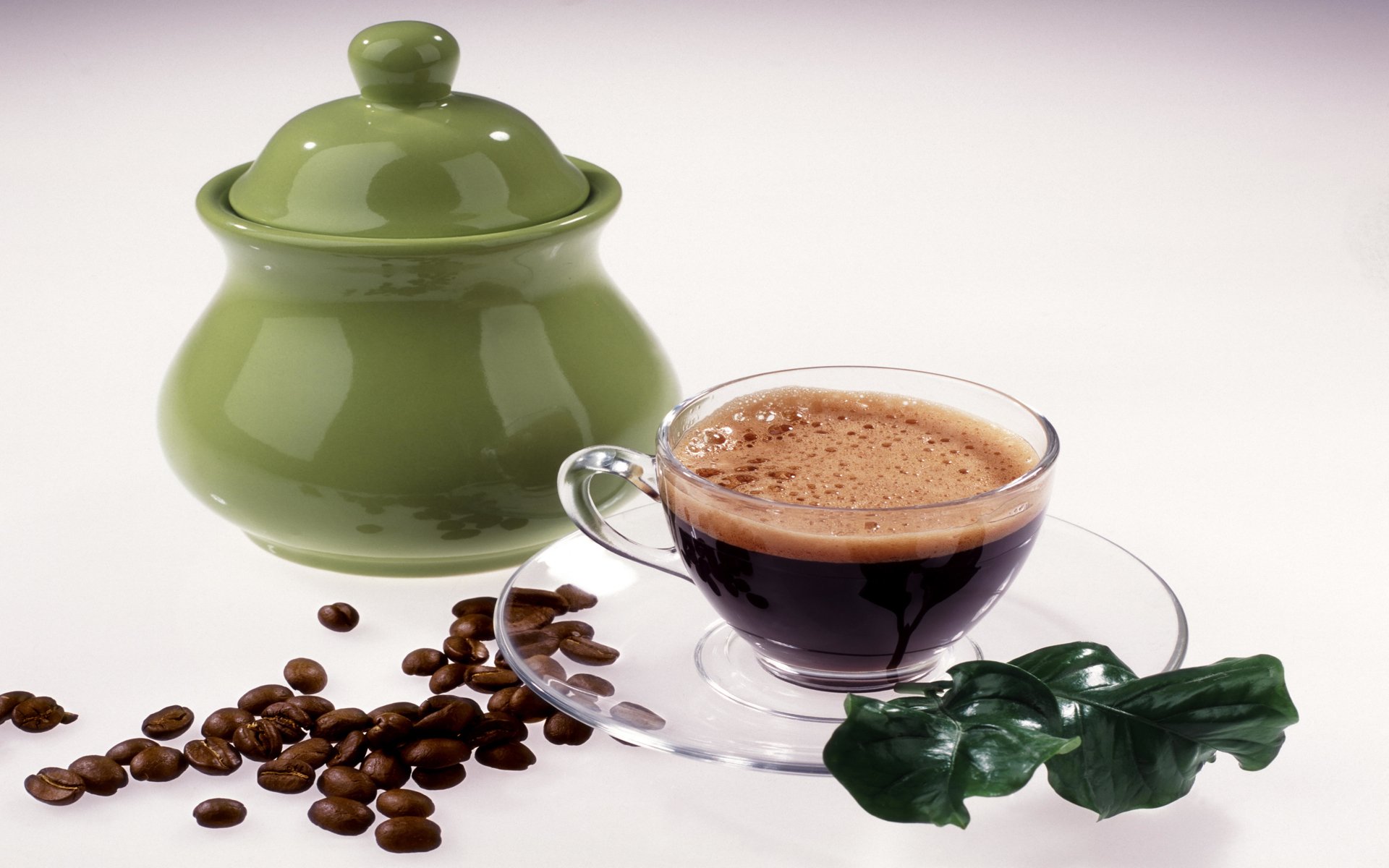 HD PC desktop wallpaper featuring a clear glass cup of espresso, scattered coffee beans, a green sugar bowl, and a dark green wrapped candy on a white background.