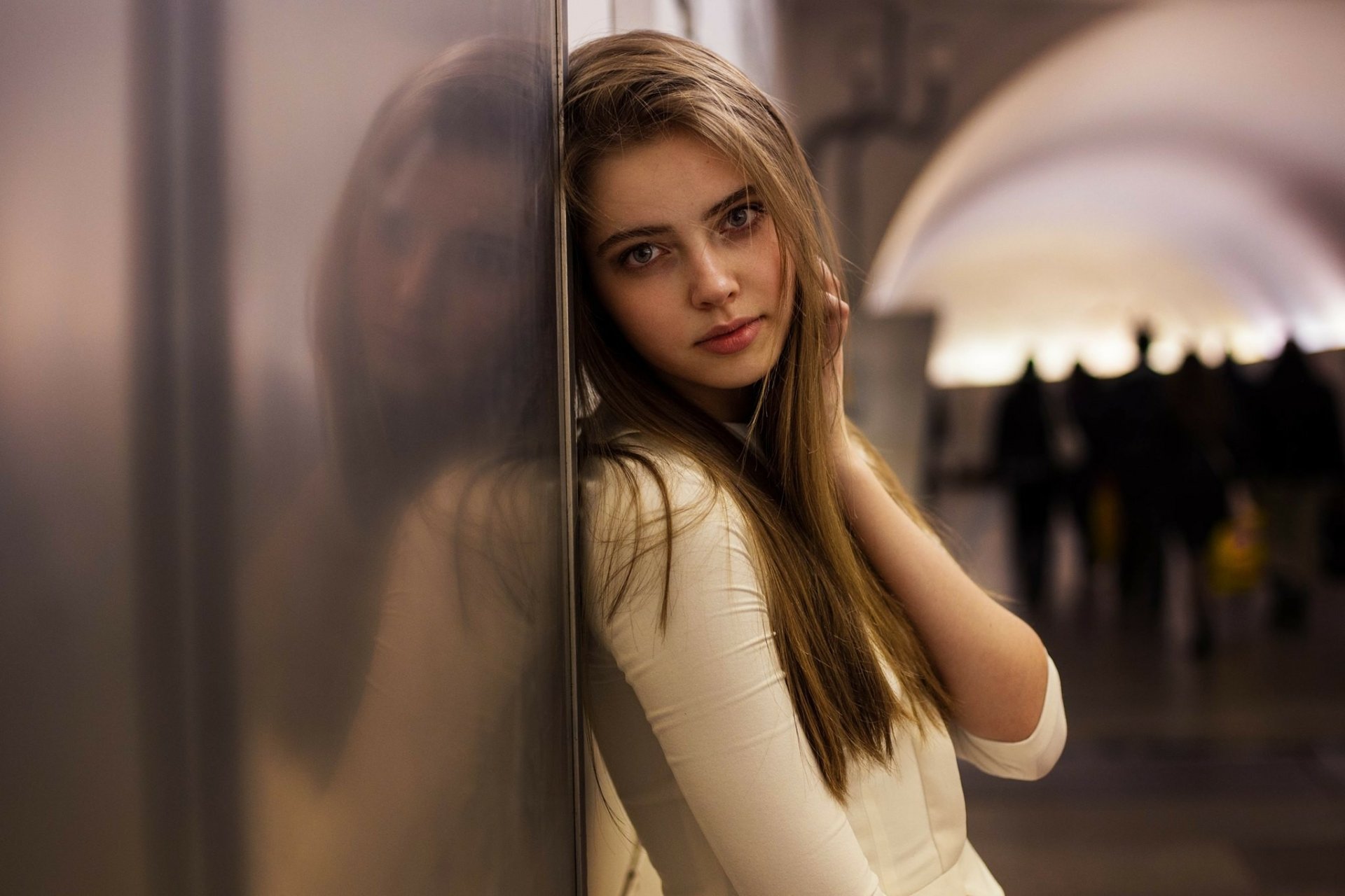 A woman with long hair leans against a reflective surface in a softly lit hallway. Image is an HD desktop wallpaper and background.