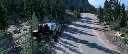 A high-definition desktop wallpaper from Furious 7, featuring a car soaring over a winding mountain road surrounded by lush trees and rugged terrain.