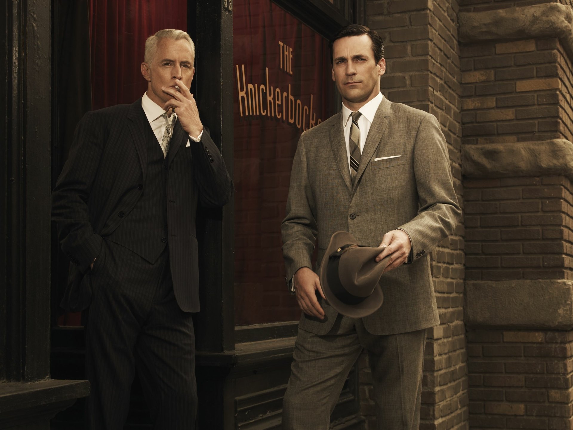 2K Quad HD Mad Men TV show desktop wallpaper: two sharply dressed 1960s men in suits stand outside an office building, one holding a hat.