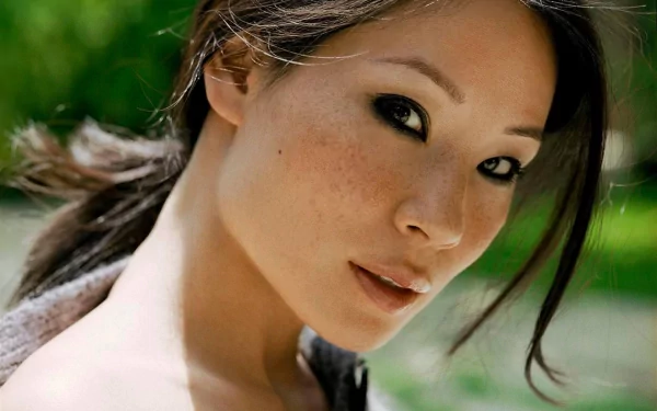 Close-up HD wallpaper of actress Lucy Liu, featuring her with soft makeup and a green, blurry outdoor background.