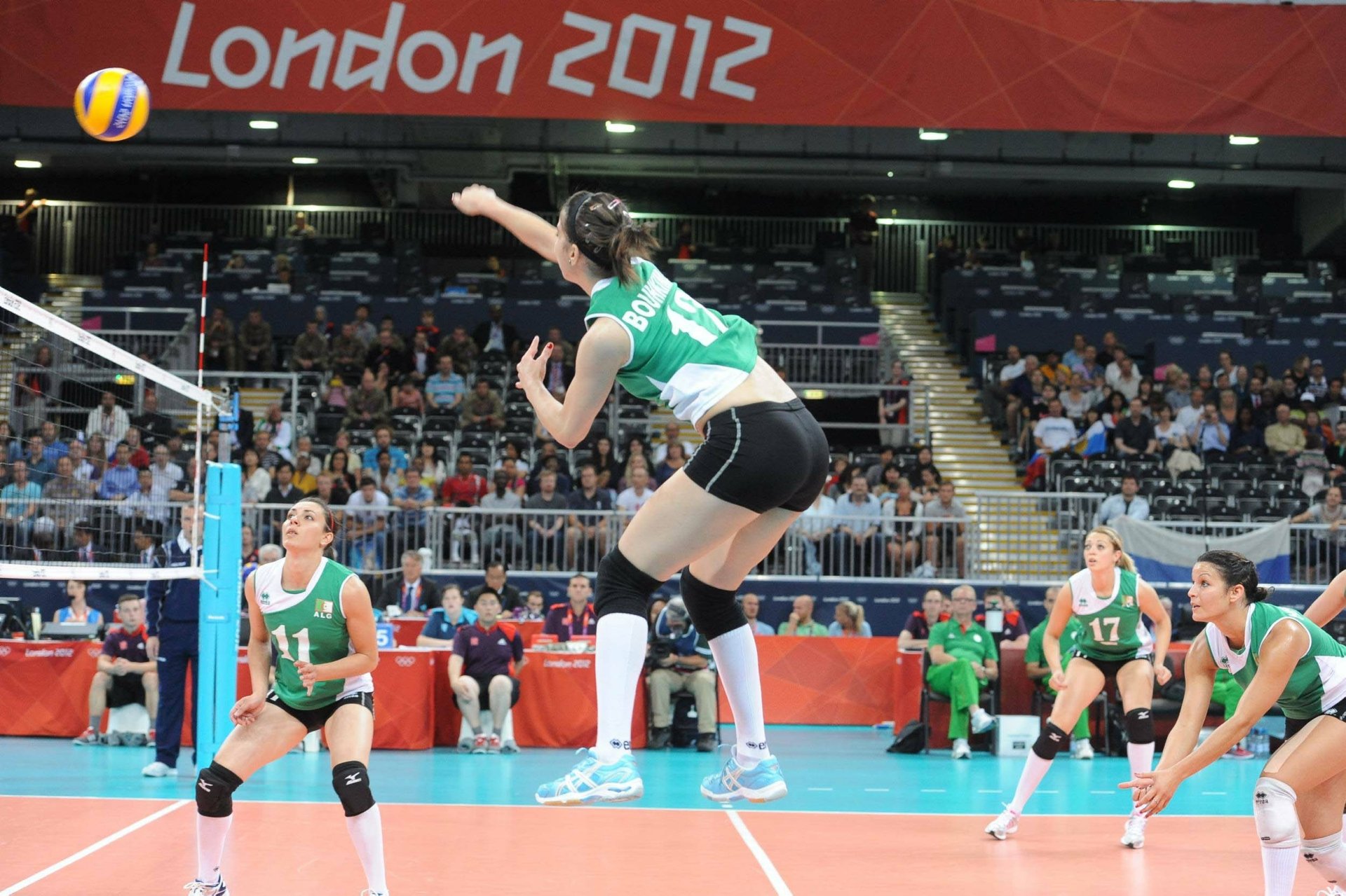 A volleyball player leaps to spike the ball during a match at the London 2012 Olympics, showcasing dynamic action in a vibrant sports setting. A stunning HD background captures the energy of the game.