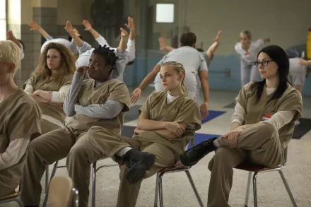 A scene from the TV show *Orange Is the New Black*, featuring characters in prison uniforms sitting in a classroom setting, showcasing their interactions and expressions. 4K Ultra HD wallpaper.