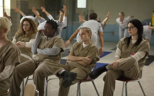 A scene from the TV show *Orange Is the New Black*, featuring characters in prison uniforms sitting in a classroom setting, showcasing their interactions and expressions. 4K Ultra HD wallpaper.