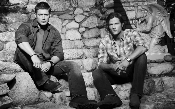 Black and white image of Sam Winchester and Dean Winchester, portrayed by Jared Padalecki and Jensen Ackles, sitting on stone steps with an angel statue in the background.