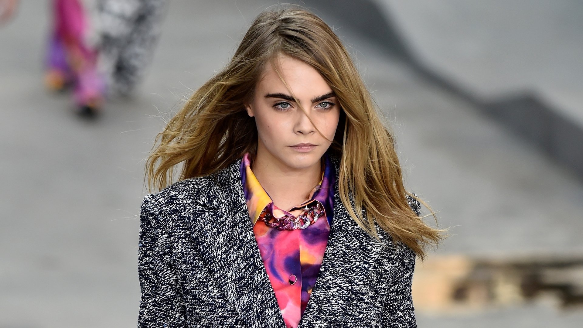 Cara Delevingne walks confidently in a bold outfit, her hair flowing in the breeze. This HD wallpaper captures her striking presence on a fashion runway.