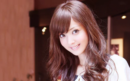 HD PC desktop wallpaper featuring celebrity Nozomi Sasaki smiling with long, wavy brown hair in a softly lit indoor setting.