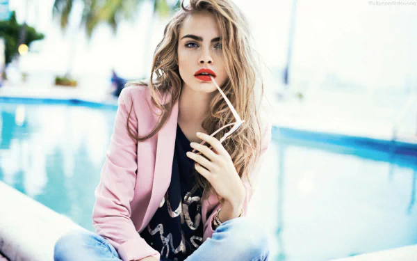 A striking HD desktop wallpaper featuring Cara Delevingne, styled elegantly with long hair, bold red lips, and a pink blazer, seated by a serene poolside backdrop.