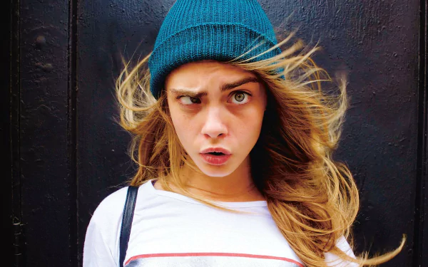 A vibrant HD desktop wallpaper featuring celebrity Cara Delevingne, showcasing her expressive face with windblown hair and a teal beanie against a dark background.