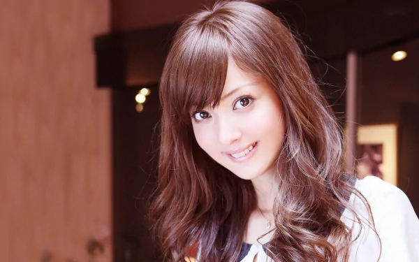 HD PC desktop wallpaper featuring celebrity Nozomi Sasaki smiling with long, wavy brown hair in a softly lit indoor setting.