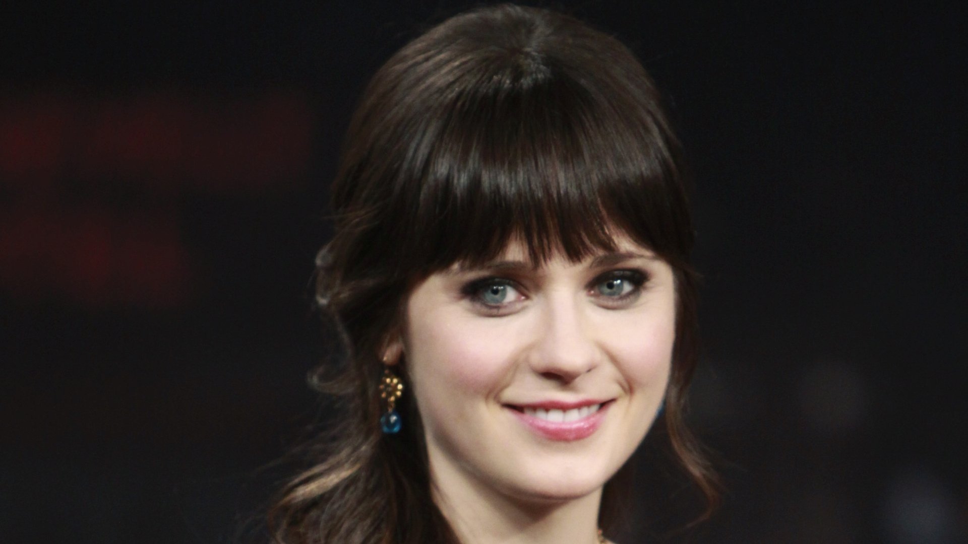HD desktop wallpaper featuring celebrity Zooey Deschanel with dark hair, bangs, and blue eyes, smiling against a blurred dark background.