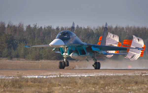 HD desktop wallpaper of a military Sukhoi Su-34 jet with deployed drag chutes on a runway, set against a backdrop of trees and a clear sky.