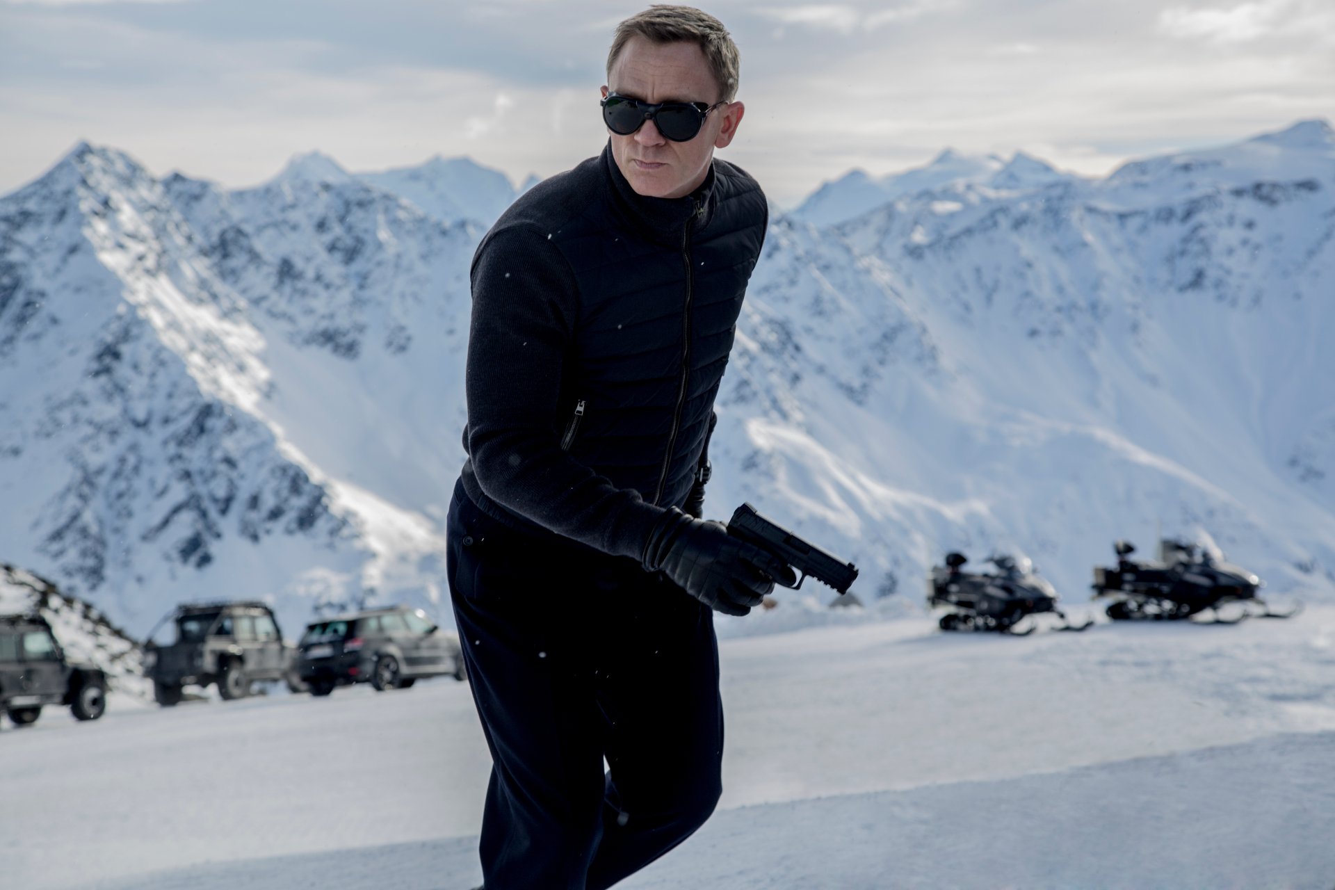 Spectre (James Bond) movie scene: armed man in black jacket and sunglasses on a snowy mountain road with vehicles in the distance — 5K Ultra HD PC desktop wallpaper background.