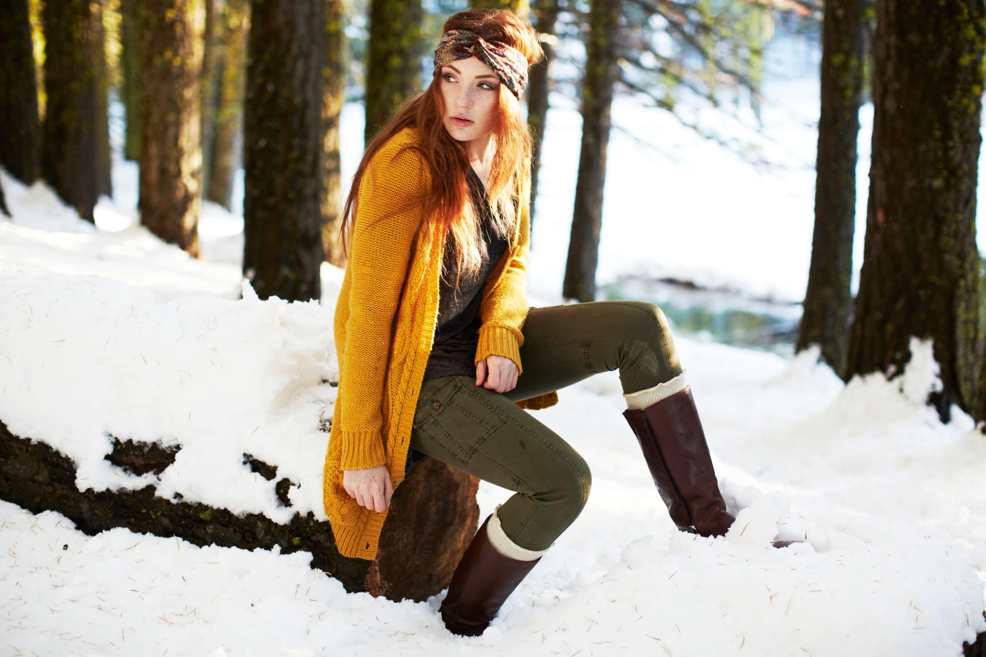 Woman in a mustard coat and headband seated on a snow-covered log in a sunlit pine forest — 5K Ultra HD PC desktop wallpaper background