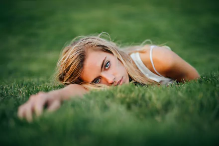 Blonde model with striking blue eyes lying in lush grass, close-up portrait — 8K Ultra HD PC desktop wallpaper/background.