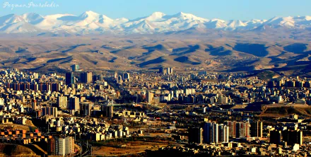 HD desktop wallpaper showcasing the man-made urban landscape of Tabriz with a backdrop of snow-capped mountains under a clear sky.