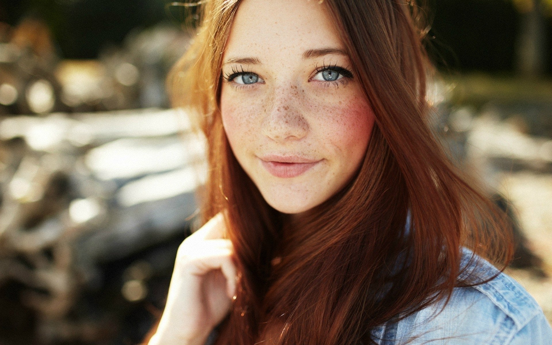HD PC desktop wallpaper featuring a close-up of a young woman with red hair, freckles, and blue eyes, softly smiling against a blurred outdoor background.
