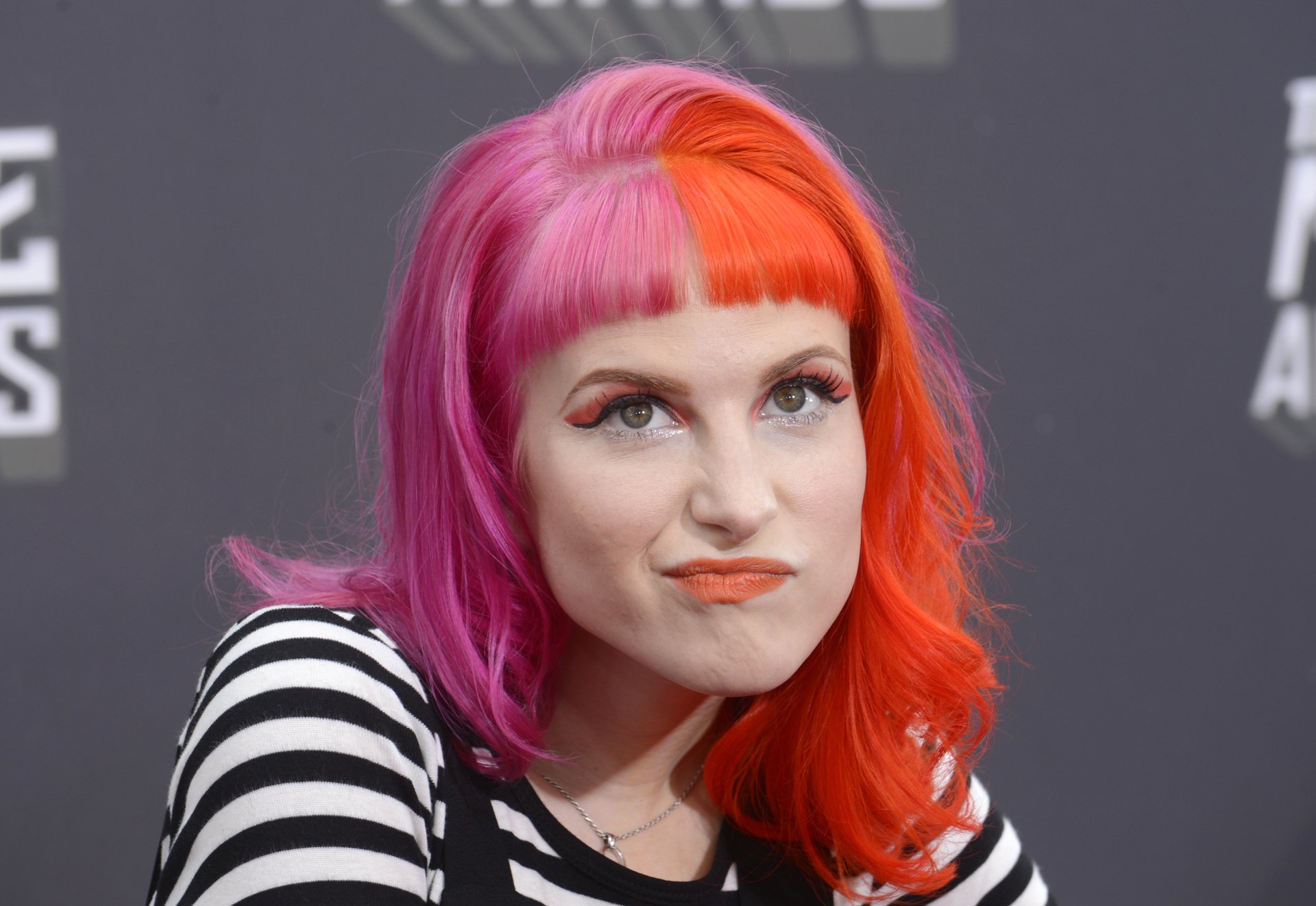 2K Quad HD PC desktop wallpaper: female music artist with half-pink, half-orange hair, bold makeup and striped top, making an expressive face against a neutral gray event backdrop.