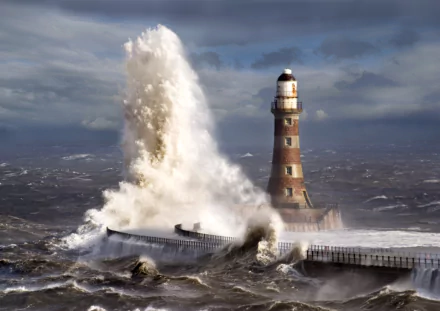 A dramatic lighthouse stands against crashing waves, with powerful water surging toward the shore, captured in stunning 8K Ultra HD for an eye-catching desktop wallpaper.
