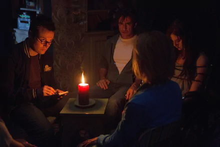 A suspenseful scene from Insidious: Chapter 3 featuring Dermot Mulroney and Stefanie Scott, where a group sits in the dark around a candle, engaged in a tense moment.