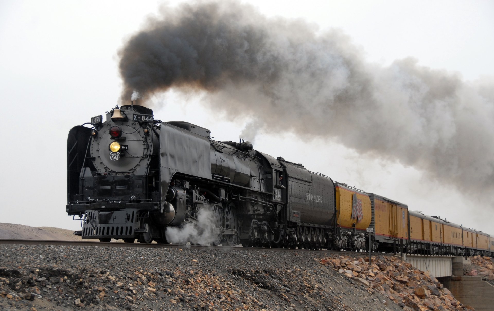 Steam locomotive pulling yellow freight cars across rocky desert, billowing smoke — 2K Quad HD PC desktop wallpaper/background.