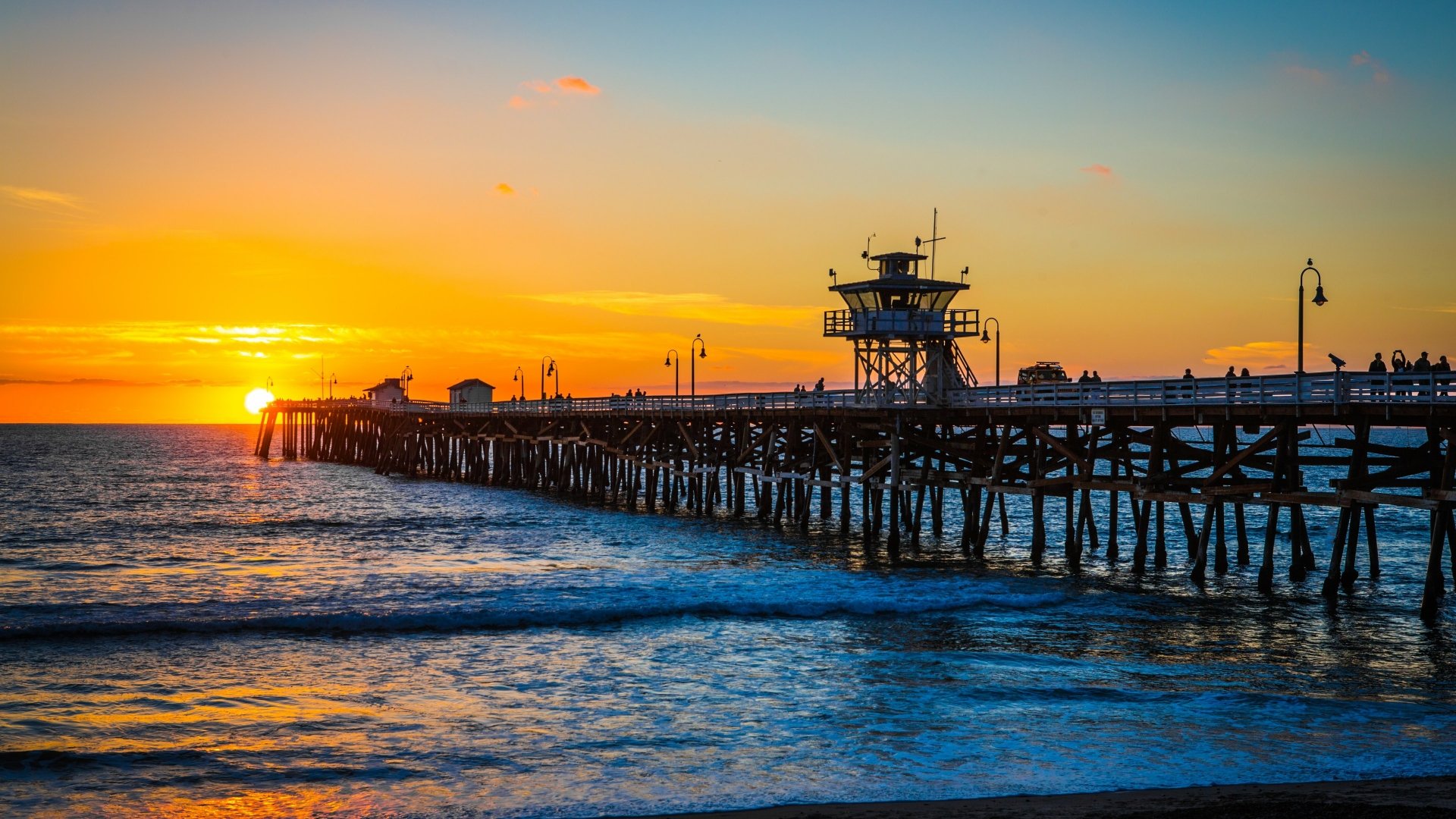 4K Ultra HD desktop wallpaper capturing a vibrant sunset over the ocean with a long man-made pier extending into the water under a colorful sky.